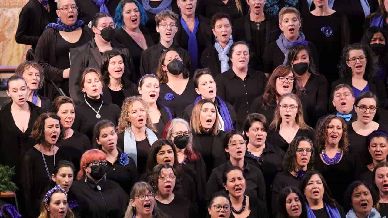 A photo SDWC singers, wearing black clothes with royal blue accents, singing in a tiered group. SDWC will peform Hope for the Holidays winter concert on December 8, 2024.